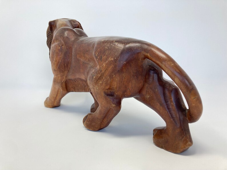May include: A hand-carved wooden lion figurine with a brown finish. The lion is depicted in a walking pose with its tail curled behind it.