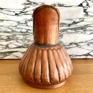 May include: A copper jug with a fluted body and a rounded spout. The jug has a hammered finish and a rustic appearance.
