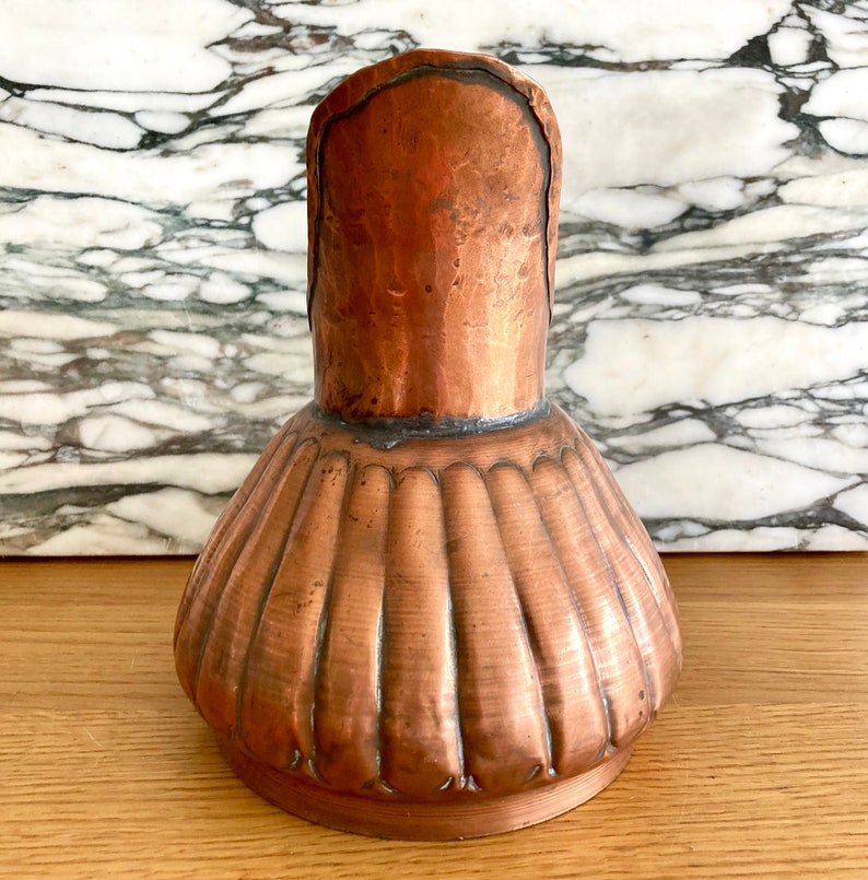 May include: A copper jug with a fluted body and a rounded spout. The jug has a hammered finish and a rustic appearance.