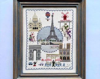 May include: Cross-stitch artwork in a wooden frame depicting Parisian landmarks. The Eiffel Tower, Arc de Triomphe, Sacré-Cœur Basilica, and Moulin Rouge are featured. The artwork includes a cafe scene and a hot air balloon, all in a detailed, colorful design.