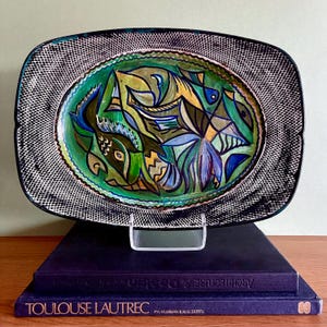 May include: A rectangular ceramic platter with a hand-painted abstract design of fish in shades of green, blue, and yellow. The platter has a black and white patterned border. The platter is displayed on a clear stand above two books.