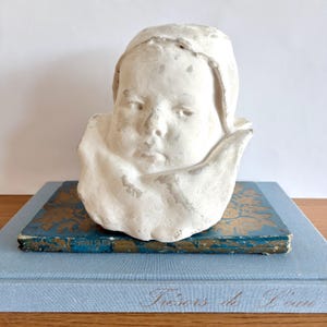 Puede incluir: Un busto de yeso blanco de la cara de un niño, con una expresión serena y un cuello drapeado. La escultura descansa sobre una pila de dos libros, el superior azul con detalles florales dorados, y el inferior azul claro con el texto "Trésors de l'eau".