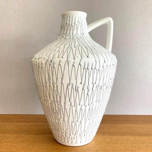 Może przedstawiać: Biały ceramiczny wazon w kształcie dzbanka z teksturowanym, wypukłym jasnoszarym wzorem pionowego zygzaka na korpusie. Naczynie posiada zwężaną szyjkę, szerokie zaokrąglone ramię oraz pojedyncze gładkie białe ucho biegnące od szyjki do ramienia.