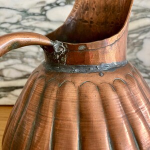 May include: A copper jug with a fluted design and a handle. The jug has a worn, aged patina and shows signs of use.