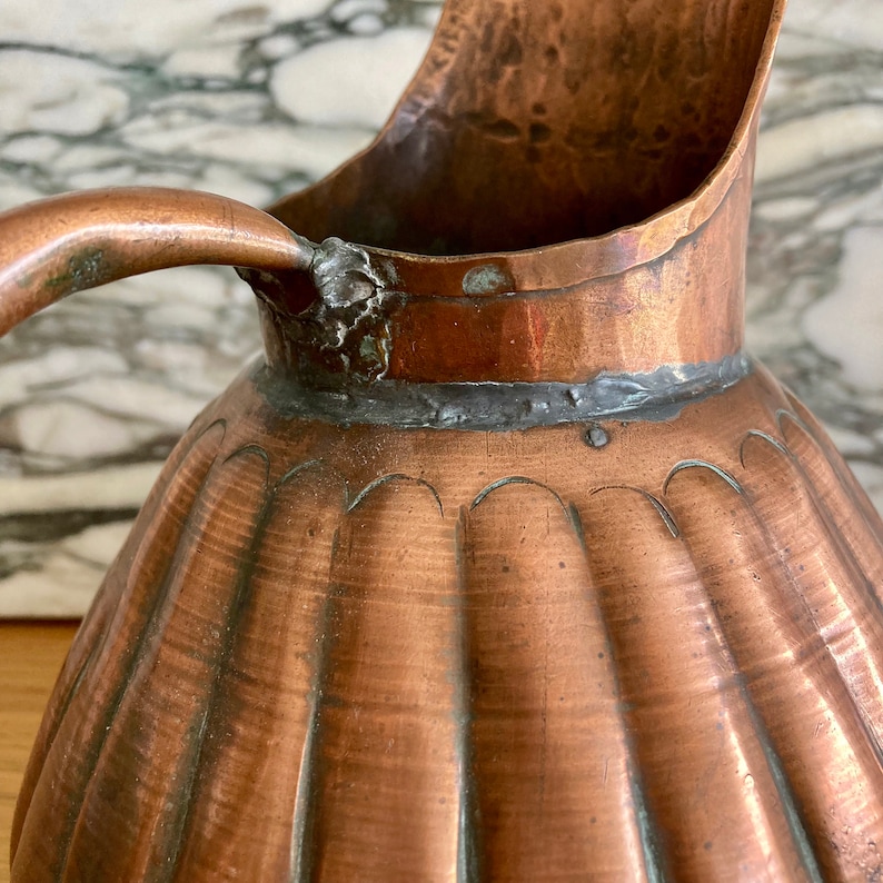 May include: A copper jug with a fluted design and a handle. The jug has a worn, aged patina and shows signs of use.