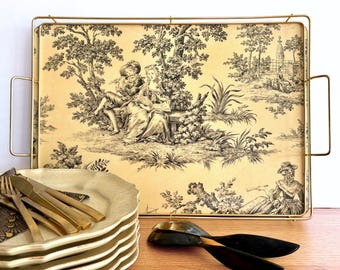 Puede incluir: Una bandeja rectangular con marco y asas de metal dorado. La superficie de la bandeja presenta una escena pastoral en blanco y negro. Una pila de platos blancos, utensilios dorados y cucharas de servir negras sobre una superficie de madera.