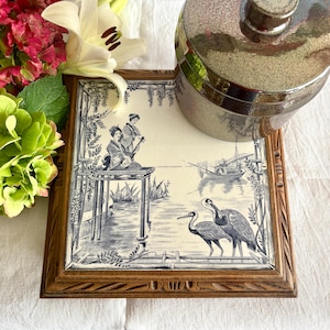 Puede incluir: Azulejo decorativo cuadrado con marco de madera con una escena de inspiración asiática azul y blanca. El azulejo representa figuras en un muelle, barcos y pájaros. Un recipiente plateado se encuentra encima, con flores en el fondo.