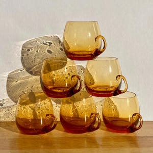 Peut inclure: Ensemble de six tasses en verre ambré avec un corps arrondi et des anses courbes. Les tasses sont empilées en forme de pyramide.