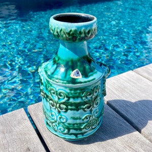 Może przedstawiać: Turkusowy i zielony ceramiczny wazon z ozdobnym wzorem. Wazon ma szeroki otwór i wąską szyjkę. Stoi na drewnianej powierzchni.