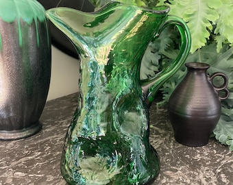 May include: A green glass pitcher with a textured surface. The pitcher has a wide mouth and a handle.