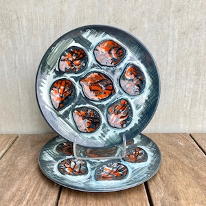May include: Two ceramic plates with a unique design. Each plate features a dark gray rim with a light blue and white interior. The interior has seven orange and black abstract shapes. The plates are displayed on a wooden surface.
