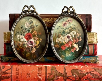 May include: Two antique oval framed floral paintings. The frames are bronze-colored with a bow at the top. The paintings feature bouquets of colorful flowers, including red, pink, and white blooms, set against a dark green background. The paintings are displayed on a stack of vintage books.
