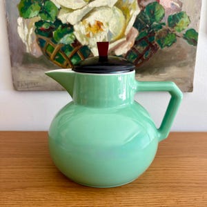 Peut inclure: Une théière en céramique vert menthe avec un couvercle noir et un bouton rouge. La théière a un corps arrondi, un bec verseur et une anse géométrique. En arrière-plan, une peinture de fleurs blanches.