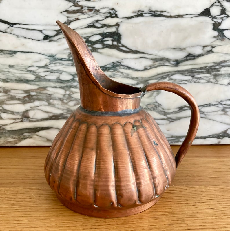 May include: A copper jug with a fluted body and a curved spout. The jug has a handle and is made of hammered copper.