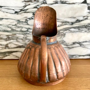 May include: A copper jug with a fluted body and a curved spout. The jug has a hammered finish and a rustic look.