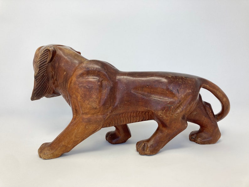 May include: A carved wooden lion figurine in a walking pose. The lion is brown and has a detailed mane. The figurine is made of dark wood and has a smooth finish.