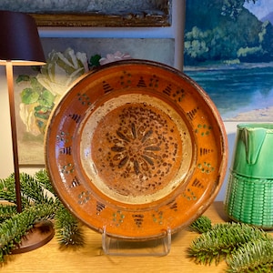 May include: A large, round, orange ceramic plate with a white center and brown and green painted designs. The plate has a raised rim and is decorated with a floral pattern in the center.