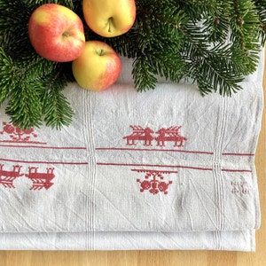 Könnte beinhalten: Eine gefaltete weiße Leinentischdecke mit roten Stickmustern von Rentieren und anderen festlichen Designs. Drei rot-gelbe Äpfel und grüne Kiefernzweige sind oben angeordnet und bilden eine saisonale Präsentation.