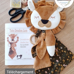 Può includere: Un giocattolo per bambini a forma di leone di peluche con una faccia marrone e bianca, circondata da una criniera marrone. Accompagnato da una coperta fantasia, un anello di legno e un modello di cucito intitolato "Tuto Doudou Lion".