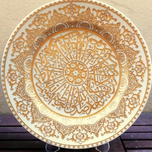 Könnte beinhalten: Ein weißer Keramikteller mit einem goldenen Dekormuster mit kunstvoller Kalligraphie. Der Teller hat einen erhöhten Rand und eine glatte Oberfläche.