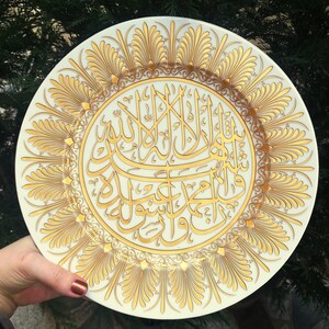Könnte beinhalten: Ein weißer Keramikteller mit goldenem Design, das komplizierte Kalligraphie in arabischer Schrift zeigt. Der Teller hat einen dekorativen Rand aus stilisierten floralen Mustern.