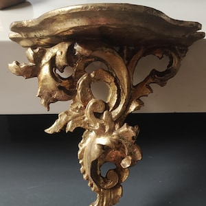 May include: A gold-colored wall shelf featuring an ornate, carved design with scrolling leaf patterns and a curved top surface. The structure has a textured, metallic finish with deep grooves and raised details throughout the entire piece.