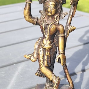 May include: A gold-colored metal statue of a deity with a crown and elaborate jewelry, standing on a circular base with decorative details. The statue is holding a flower in one hand and has a serene expression.