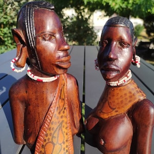 Puede incluir: Dos esculturas de madera de personas con detalles intrincados. Las esculturas están talladas en madera oscura y pintadas con acentos naranja, rojo y blanco. Llevan collares y pendientes de cuentas.