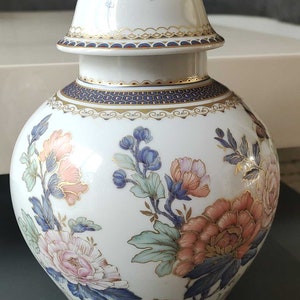 May include: A white porcelain vase with a gold lid and floral designs in shades of pink, blue, and green. The vase has a blue and gold trim around the top and bottom.