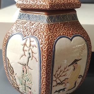 May include: A decorative ceramic jar with a lid, in brown and white. The jar has a detailed pattern on the lid and sides, with illustrations of figures in a landscape. The lid has a blue finial. The jar is likely of Asian origin.