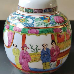 Könnte beinhalten: Eine weiße Porzellan-Dose mit Deckel, verziert mit einem bunten floralen und geometrischen Muster. Die Dose zeigt eine Szene mit Menschen in traditioneller chinesischer Kleidung.