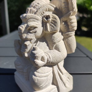 Peut inclure: Statue en pierre beige d'une divinité assise avec plusieurs bras et une coiffe ornée. La statue présente des traits de visage détaillés et tient un petit objet. La sculpture est sur une base carrée et a une surface texturée.