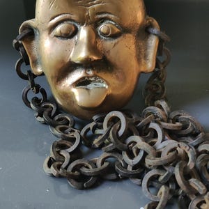 May include: A brass mask with a chain attached. The mask depicts a face with a serious expression. The chain is made of metal links.
