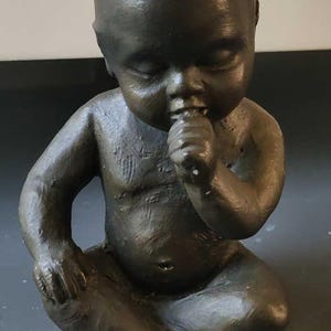 May include: A bronze-colored sculpture of a baby in a seated position, with its eyes closed and a finger in its mouth. The baby's body is detailed, showing the folds of skin and the shape of the limbs. The sculpture is on a dark surface.