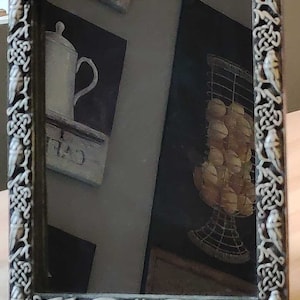 Puede incluir: Un espejo negro enmarcado con un diseño de nudo celta. El espejo refleja una jarra blanca y una cesta de alambre llena de objetos redondos.