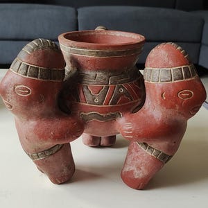 以下が含まれることがあります： 2つの様式化された人物が持つ赤い陶器の容器。人物と容器は幾何学模様で装飾されており、赤茶色です。この作品はおそらく装飾品または儀式用のアイテムです。