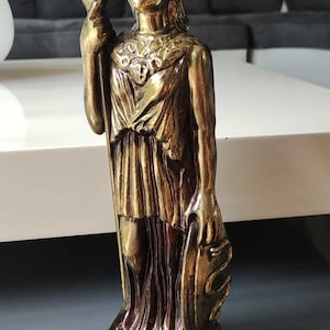 Puede incluir: Una estatua dorada de Atenea, la diosa griega de la sabiduría y la guerra, sosteniendo una lanza y un escudo. La estatua está de pie sobre una base con la palabra "ATHINA" inscrita en ella.
