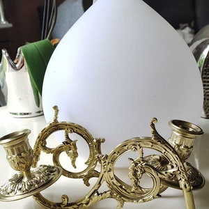 Könnte beinhalten: Verzierter Messing-Kerzenhalter mit zwei Kerzenschalen und dekorativen Verzierungen. Der Kerzenhalter hat ein detailliertes Design mit floralen und Blattmotiven. Im Hintergrund befindet sich ein weißes, tropfenförmiges Objekt.