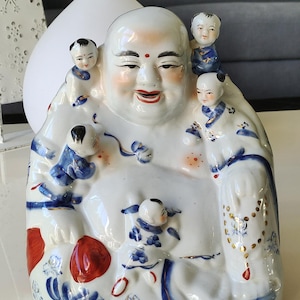 May include: A white ceramic Buddha figurine with a smiling face and small children climbing on it. The figurine has blue and red accents, with a gold-colored necklace. The Buddha is a decorative item.