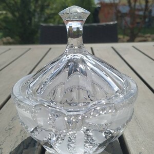 Peut inclure: Un pot à bonbons en verre transparent avec un design décoratif et orné. Le pot a un couvercle avec un sommet pointu.