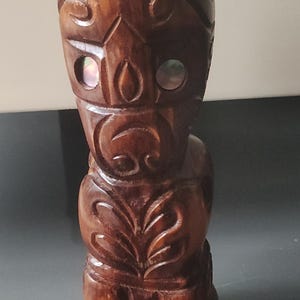 Könnte beinhalten: Eine geschnitzte Tiki-Statue aus Holz mit dunkelbraunem Finish. Die Statue weist detaillierte Schnitzereien von Gesichtszügen, Mustern und einem Blattdesign auf dem unteren Teil auf. Kleine, schillernde Muscheleinlagen sind sichtbar.