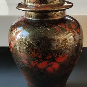 Peut inclure: Un vase en céramique rouge et or avec un couvercle. Le vase a un motif marbré avec des accents dorés.