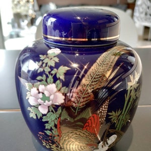 Op de afbeelding: Een kobaltblauwe keramische pot met een gouden deksel en een gedetailleerd bloemen- en vogelontwerp. De pot heeft een fazant met gouden en rode accenten, omringd door bloemen en bamboe.