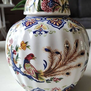 May include: A white ceramic jar with a lid, decorated with colourful floral and bird designs. The jar features a peacock with a long tail, a red bird, and blue and yellow flowers.