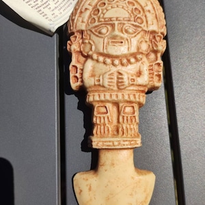 May include: A beige ceramic figurine of a deity with a human-like face and a headdress. The figurine is wearing a necklace and has a skirt-like garment. The figurine is holding its hands together in front of its chest. The figurine is standing on a flat base.