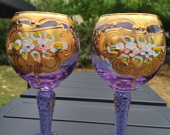 May include: Two purple glass wine goblets with gold and white floral designs. The goblets have a textured stem and a round base.