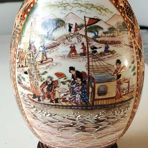 Peut inclure: Un vase en porcelaine blanche en forme d'œuf avec une scène peinte à la main détaillée de personnes dans des bateaux sur une rivière. Le vase est orné d'une bordure dorée et repose sur un socle en bois foncé.