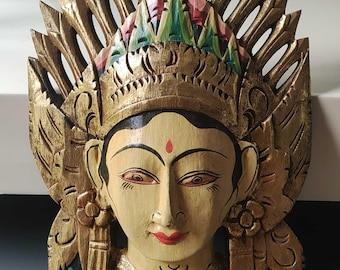 May include: A wooden wall hanging mask with a gold, green, and red painted design. The mask depicts a woman's face with a crown and intricate details.
