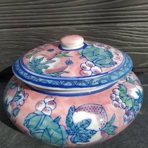 Peut inclure: Un pot en céramique rose et bleu avec un couvercle, décoré d'un motif floral de feuilles vertes, de fleurs roses et de baies blanches. Le pot a une forme ronde et une surface lisse.