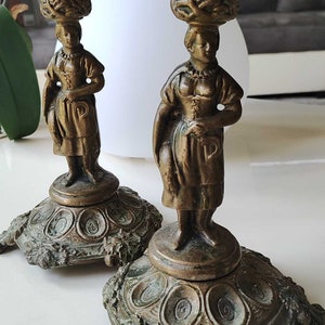 Puede incluir: Un par de candelabros antiguos de latón que representan a una mujer con un vestido tradicional sosteniendo una jarra. Los candelabros tienen una base detallada con un diseño floral.